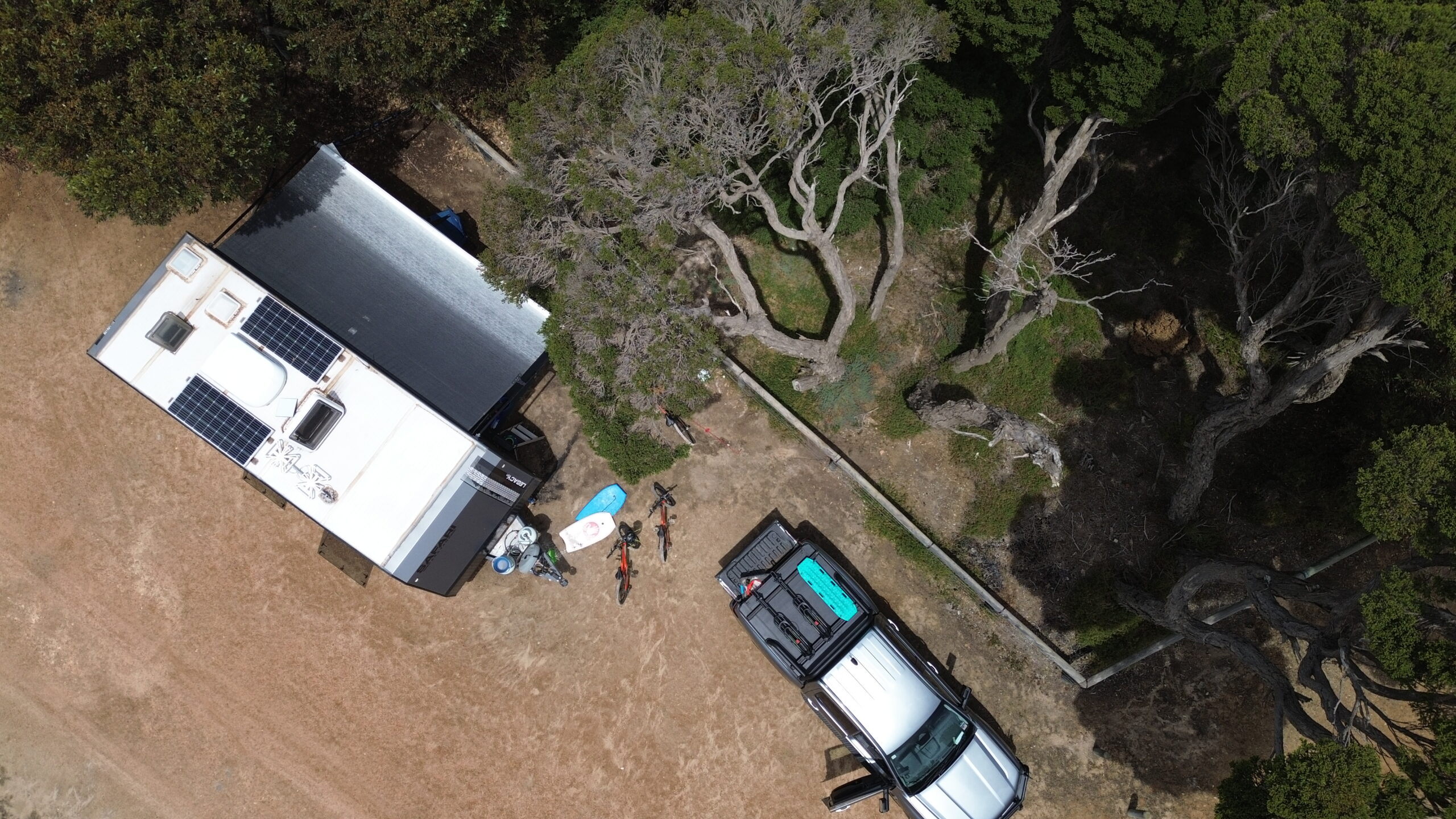 Overhead drone shot of a caravan and a 4WD parked side by side on dry grass beside gnarled coastal trees, with bikes, a paddleboard and camp gear laid out between them